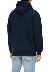 Hombre vistiendo una sudadera oscura azul marino y jeans claros, visto desde atrás contra un fondo blanco.