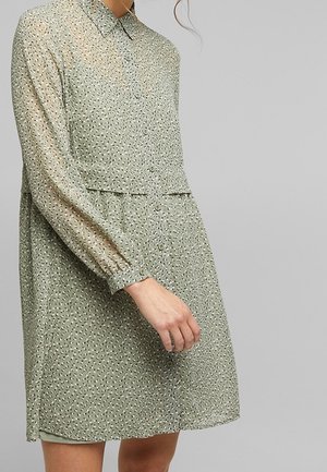 Transparante groene jurk met een knoopsluiting aan de voorkant, een kraag en lange mouwen. Voorzien van kleine, contrasterende zwarte bloemmotieven door het hele ontwerp.