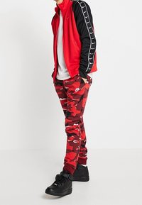 Röd och svart träningsoverall med en zipjacka, som har vita accenter och byxor med kamouflage-mönster. Bärs med svarta sneakers.