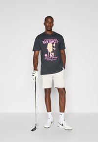 Svart grafisk T-skjorte med rosa tekst og en tegneseriemarv karakter av en pølse. Kombinert med beige shorts, hvite sokker og joggesko, holder en golfkølle.