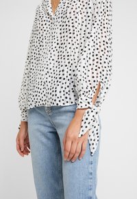 White sheer blouse with black polka dots, featuring a V-neck, button closure, and tied cuffs. Paired with light blue jeans.