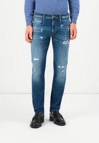 Meeste kulunud sinised teksad, mis on valmistatud denimist, slim fit lõikega, rebenemistega ja pleekinud viimistlusega. Kombineeritud mustade slip-on kingadega.