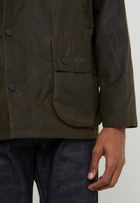 Olive green waxed jacket with a buttoned front, large patch pockets, subtle branding, and a relaxed fit. Smooth texture, mid-length design.