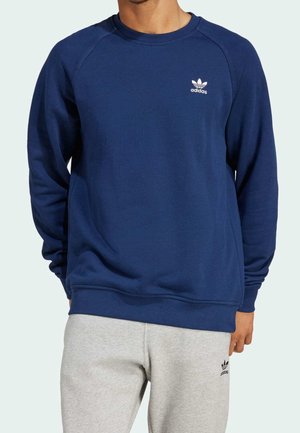 Homme portant un sweat-shirt Adidas bleu marine et un pantalon de survêtement Adidas gris clair, visible du cou jusqu'au milieu de la cuisse sur un fond uni.