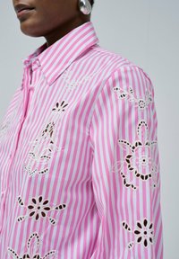 Chemise rayée rose et blanche avec broderies florales et découpes. Comprend un col et une fermeture à boutons, fabriquée en tissu léger.