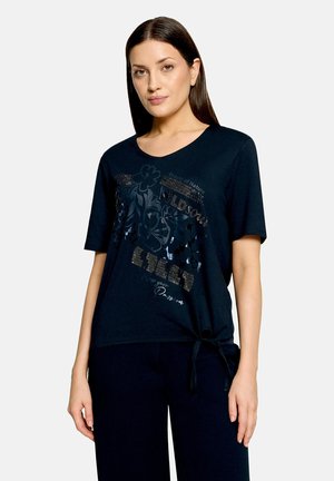 Woman with straight dark hair wearing a black short-sleeve top with floral print and text, tied at one side, standing against a plain background.