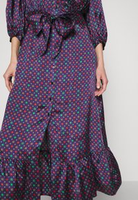 Robe bleue avec motif d’étoiles rouges et vertes et motifs floraux, boutonnée sur le devant, ourlet à volants et nœud noué à la taille, portée par une personne aux bras détendus.