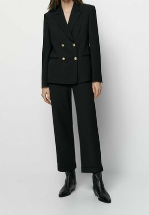 Woman wearing a black double-breasted blazer with gold buttons, matching wide-leg trousers, and black ankle boots against a plain background.