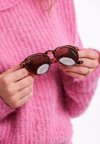 Kočičí sluneční brýle ve tvaru želvoviny s hnědými skly, světlými akcenty a lesklým povrchem, držené v rukou oblečených v texturovaném růžovém svetru.