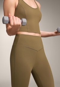 Osoba ubrana w oliwkowozielony top i legginsy do ćwiczeń, trzymająca małe szare hantle w trakcie ćwiczeń z zgiętymi ramionami, na jasnym neutralnym tle.