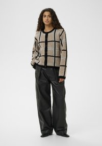 Bežs un melns raibs kardigans ar pogām, kas izceļas ar mīkstu tekstūru un garām piedurknēm, apvienots ar platām melnām mākslīgās ādas biksēm.