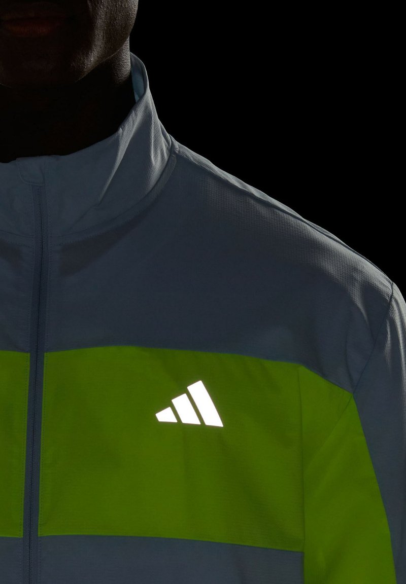 Hellblau und grüne Sportjacke mit reflektierendem Adidas-Logo, durchgehendem Reißverschluss, hohem Kragen und strukturiertem Stoff. Dunkler Hintergrund verbessert die Sichtbarkeit.