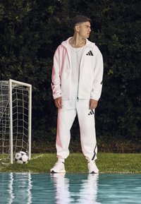 White athletic tracksuit with black accents, featuring a hooded jacket and joggers. Standing by a soccer goal with a ball nearby.
