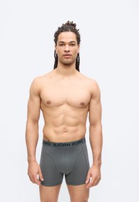 Homme musclé aux dreadlocks, debout face à l'avant, portant un boxer gris Björn Borg sur un fond blanc uni.