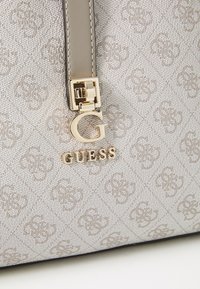 Beige Guess håndveske med en teksturert overflate og et subtilt logomønster. Gullhardware og et fremtredende logoaccent ved glidelåsen.