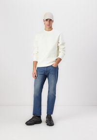 Sudadera beige claro con logo en relieve, jeans rectos azules y zapatillas gris oscuro. El modelo lleva una gorra beige a juego.