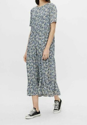 Nainen, joka pukeutuu sinivalkoiseen kukkakuvioiseen midimekkoon, jossa on lyhyet hihat, yhdistettynä mustavalkoisiin tennareihin, seisoo valkoista taustaa vasten.