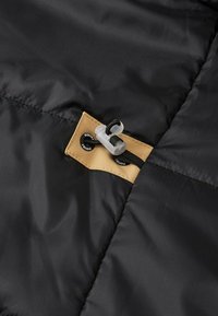 Sezione di giacca a vento nera che mostra un'etichetta di tessuto color sabbia con un cordino e due bottoni neri, caratterizzata da una texture trapuntata e una finitura liscia.