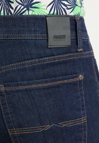 Ciemnoniebieskie dżinsy o teksturowanym wykończeniu, z pomarańczowymi przeszyciami i czarną skórzaną naszywką z napisem "PIONEER AUTHENTIC JEANS".