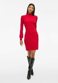 Robe tricotée rouge avec un col haut et de longues manches bouffantes, silhouette ajustée, associée à des bottes noires montantes à talons pointus.