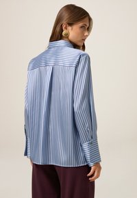 Blaue gestreifte Bluse mit langen Ärmeln, die auf der Rückseite einen Faltenwurf und Knopfdetails an den Ärmelbündchen aufweist. Glattes Material mit sanftem Glanz.