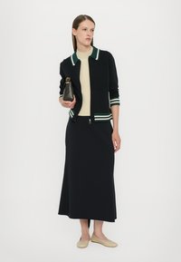 Cardigan negru din tricot cu guler în dungi verzi și albe, asortat cu o fustă neagră care se încadrează pe corp și balerini bej; ținând o geantă mică neagră.