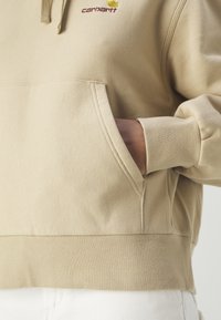 Beige hoodie made from soft fabric, featuring a kangaroo pocket, ribbed cuffs, and a subtle embroidered logo on the chest.