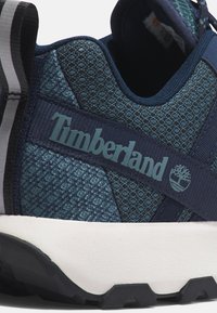 Zapatilla deportiva azul oscuro con parte superior de malla texturizada, acentos reforzados y un prominente logo de Timberland. Suela de goma blanca, diseñada para la comodidad.