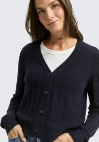 Marineblauer Cardigan mit gerippter Textur, V-Ausschnitt und fünf vorderen Knöpfen. Lange Ärmel mit schmalem Bund. Über einem weißen Oberteil getragen.