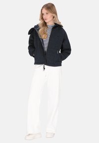 Jachetă texturată albastru închis cu glugă și fermoar, purtată peste un top cu dungi, asortată cu pantaloni largi albi și pantofi deschiși la culoare.