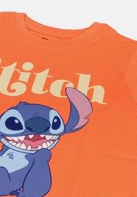 Oranssi puuvillapaita, jossa on graafinen kuva Stitchistä, sinisestä hahmosta, jolla on suuret silmät, hymyillen sanan "Stitch" vieressä kermavärisellä kirjasimella.