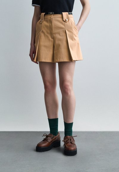 Personne portant un short plissé couleur chameau avec des bretelles à boutons, des chaussettes vert foncé et des chaussures à plateforme marron à lacets, debout sur un sol gris.