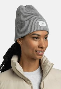 Gorro de lana gris con una textura acanalada y un dobladillo doblado, que presenta una pequeña etiqueta blanca. Llevado con una chaqueta de plumas beige.
