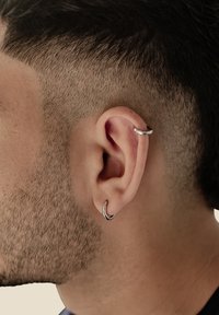 Pendientes de aro de plata adornan una oreja cortada al ras, exhibiendo un acabado metálico liso y pulido. El peinado presenta los lados cortos y desvanecidos.