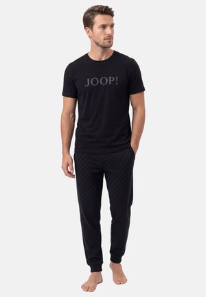 Mand iført en sort "JOOP!" t-shirt og sorte joggebukser med mønster, stående barfodet med den ene hånd i lommen, kigger mod højre.