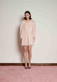 Kvinna står mot en slät vägg iklädd en rosa texturerad miniklänning med långa ärmar och leopardmönstrade högklackade skor.