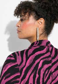 Blazer a righe zebrate rosa e nere con una texture liscia, caratterizzato da un colletto a punta e una vestibilità sartoriale. Orecchini lunghi e lucidi aggiungono stile.