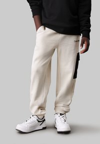 Crème sweatpants met een zwarte patch pocket en Calvin Klein-logo. Gecombineerd met witte sneakers met zwarte accenten. Zacht, textuurstof.