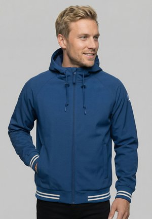 Hombre rubio con una chaqueta azul con capucha y cremallera, con puños y dobladillo con rayas blancas, manos en los bolsillos, mirando a la derecha con una leve sonrisa.