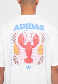 Balta marškinėlių su omaro piešiniu, apsuptu citrinų riekelių, rožinės spalvos čekiu raštu ir mėlynu Adidas logotipu.