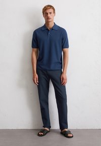 Marineblauwe polo met korte mouwen, gecombineerd met donkerblauwe broek en zwarte sandalen. Gladde textuur en klassiek kraagontwerp.