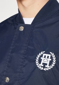 Navy bomber jacket with a ribbed collar, front snap buttons, and white embroidered logo inside a laurel wreath on the left chest.