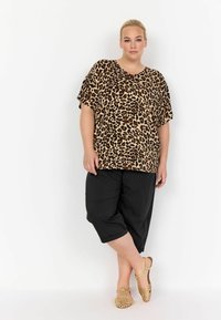 Top s leopardím potiskem a krátkým rukávem, v kombinaci s černými zkrácenými kalhotami. Modelka má na sobě světle hnědé sandály na plochém podpatku s vykrojeným vzorem. Hladký materiál.
