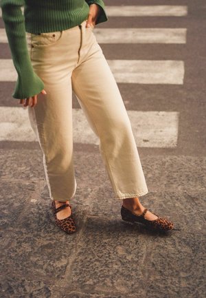 Ballerine con stampa leopardata e cinturino, indossate con jeans bianchi a gamba larga e un maglione verde a costine, su un pavimento in pietra texturizzata.