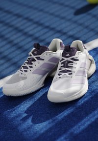 Chaussures de sport blanches et violettes avec lacets reposant sur un terrain de sport bleu près d'une ligne blanche.