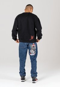 Black sweatshirt with a crew neck and logo detail; blue jeans with a colorful embroidered skull and eagle design on the back. Black sneakers.