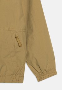 Giacca leggera beige con tessuto texturizzato, polsini elasticizzati e una tasca con zip dotata di un anello per il tirante.