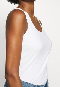 Vit ribbad tanktop med rundad halsringning, tillverkad av stretchtyg. Bärs med högmidjade jeans. Slät textur, åtsittande design.