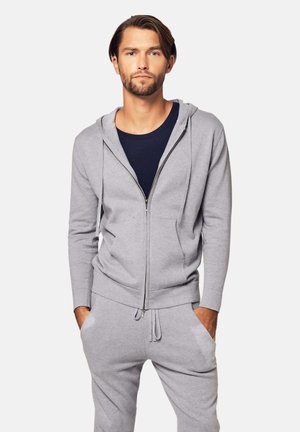 Mann mit Bart, der einen grauen Kapuzenpullover mit Reißverschluss und passende Jogginghose trägt, steht mit den Händen in den Taschen vor einem einfarbigen Hintergrund.