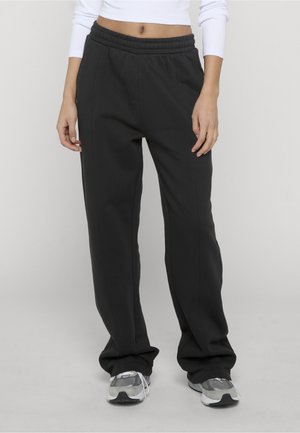 Personne portant un pantalon noir à taille élastique et des baskets grises, debout devant un fond clair uni.